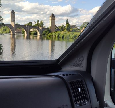 Foto a través del vidre de davant de La Camper amb un riu i una pont antic amb dues torres al fons.